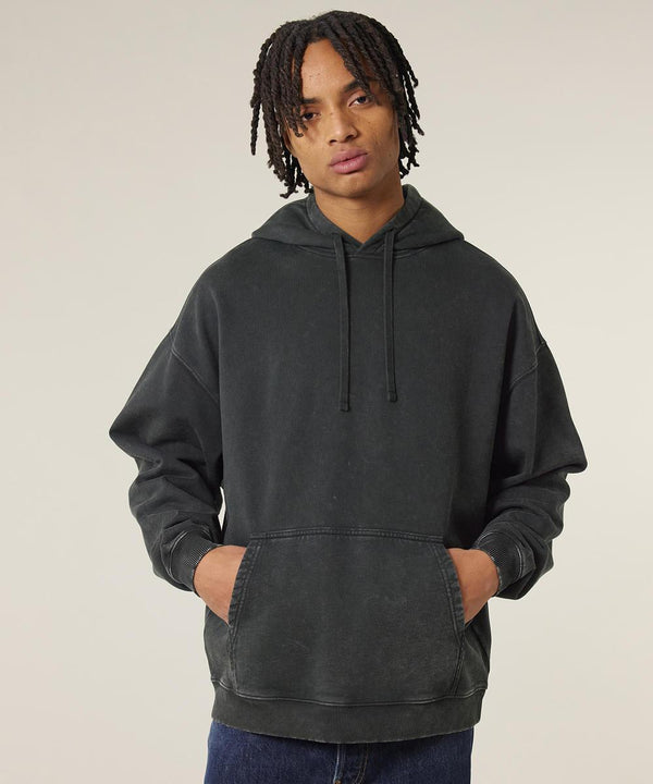 Wearing the Slammer 2.0 Vintage (STSU211) by Stanley/Stella, a person with medium-length dreadlocks stands against a light background in a dark grey hooded sweatshirt and blue jeans, hands in the front pocket.