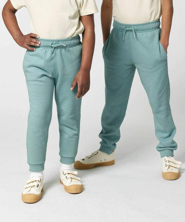 Two children standing, each wearing light-colored shirts, teal sweatpants, and white sneakers SX716 by Stanley/Stella. Hands in pockets and arms relaxed. Plain white background.