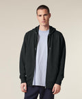 A person wearing the Stanley/Stella Connector 2.0 (STSU207) black zip-up hoodie over a light grey T-shirt and dark trousers stands against a plain, light-coloured background.