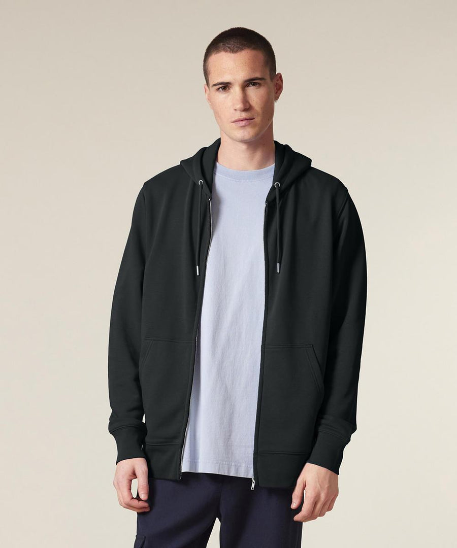 A person wearing the Stanley/Stella Connector 2.0 (STSU207) black zip-up hoodie over a light grey T-shirt and dark trousers stands against a plain, light-coloured background.