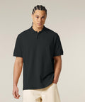 A person with plaited hair wears the Stanley/Stella Prepster 2.0 (STPU222) dark short-sleeve polo and light trousers, standing against a plain light background with one hand in their pocket.