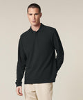 A man stands against a plain light background, wearing the Stanley/Stella Prepster 2.0 long sleeve (STPU223) in black with beige trousers, facing forward with a neutral expression and one hand relaxed by his side.