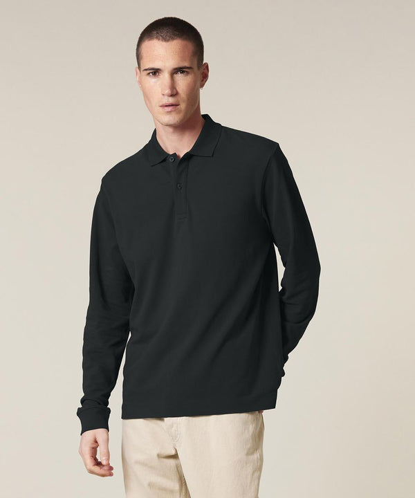 A man stands against a plain light background, wearing the Stanley/Stella Prepster 2.0 long sleeve (STPU223) in black with beige trousers, facing forward with a neutral expression and one hand relaxed by his side.