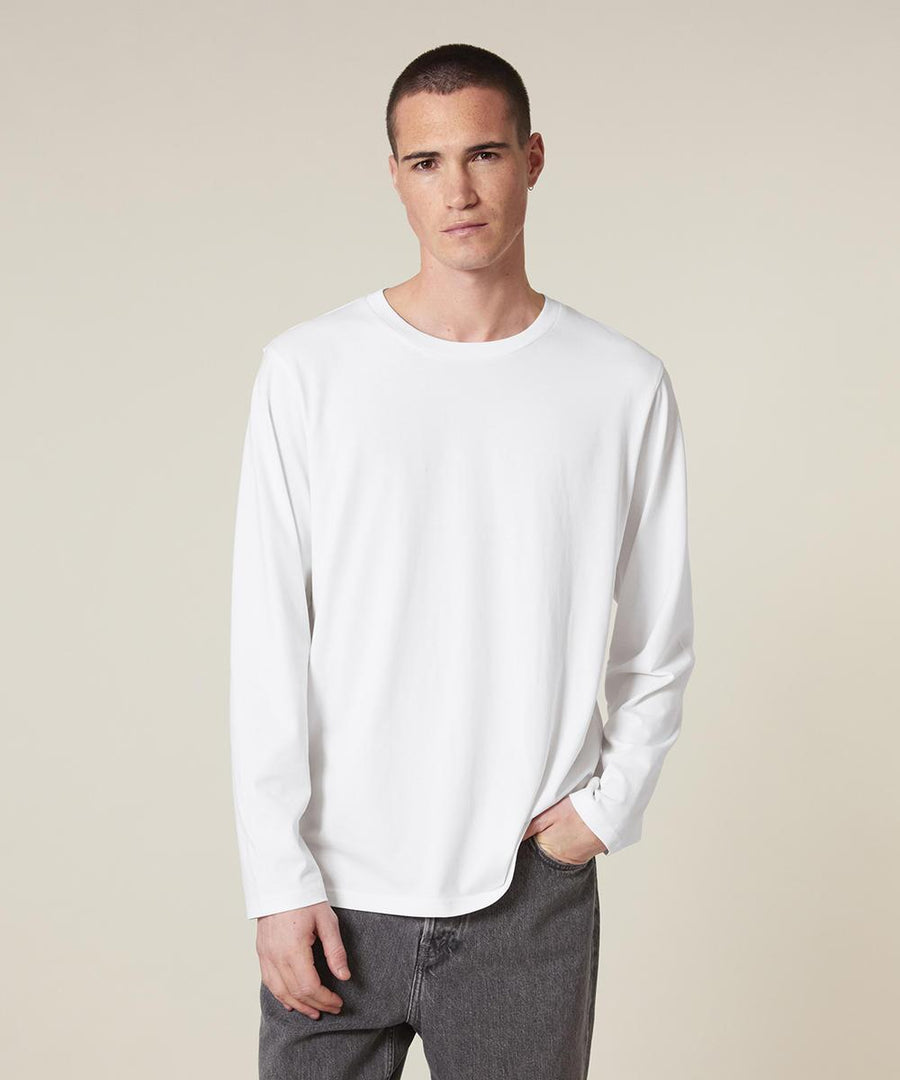 A person with short dark hair wears the Stanley/Stella Creator 2.0 long sleeve (STTU199) and dark grey jeans, standing against an off-white background with one hand in their pocket.