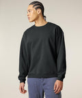 A person with plaited hair wears a loose-fitting Stanley/Stella Radder 2.0 Vintage (STSU210) black sweatshirt and blue trousers, standing against a plain light background and looking to their left.