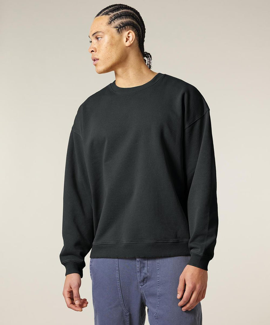 A person with plaited hair wears a loose-fitting Stanley/Stella Radder 2.0 Vintage (STSU210) black sweatshirt and blue trousers, standing against a plain light background and looking to their left.