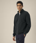 A man stands against a plain background, wearing the Stanley/Stella Trailer 2.0 (STSU253) black zip-up jacket over a white shirt and dark trousers. He looks forward neutrally, with one hand by his side.