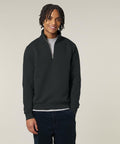 A person stands against a plain background, smiling with one hand by their side. They wear the Stanley/Stella Trucker 2.0 (STSU204) black quarter-zip jumper over a light grey shirt and dark trousers, with shoulder-length twisted hair.