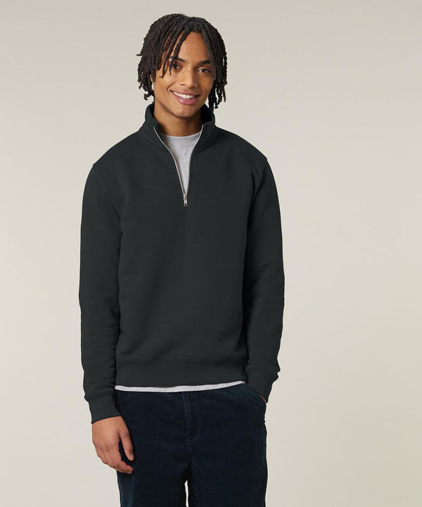 A person stands against a plain background, smiling with one hand by their side. They wear the Stanley/Stella Trucker 2.0 (STSU204) black quarter-zip jumper over a light grey shirt and dark trousers, with shoulder-length twisted hair.