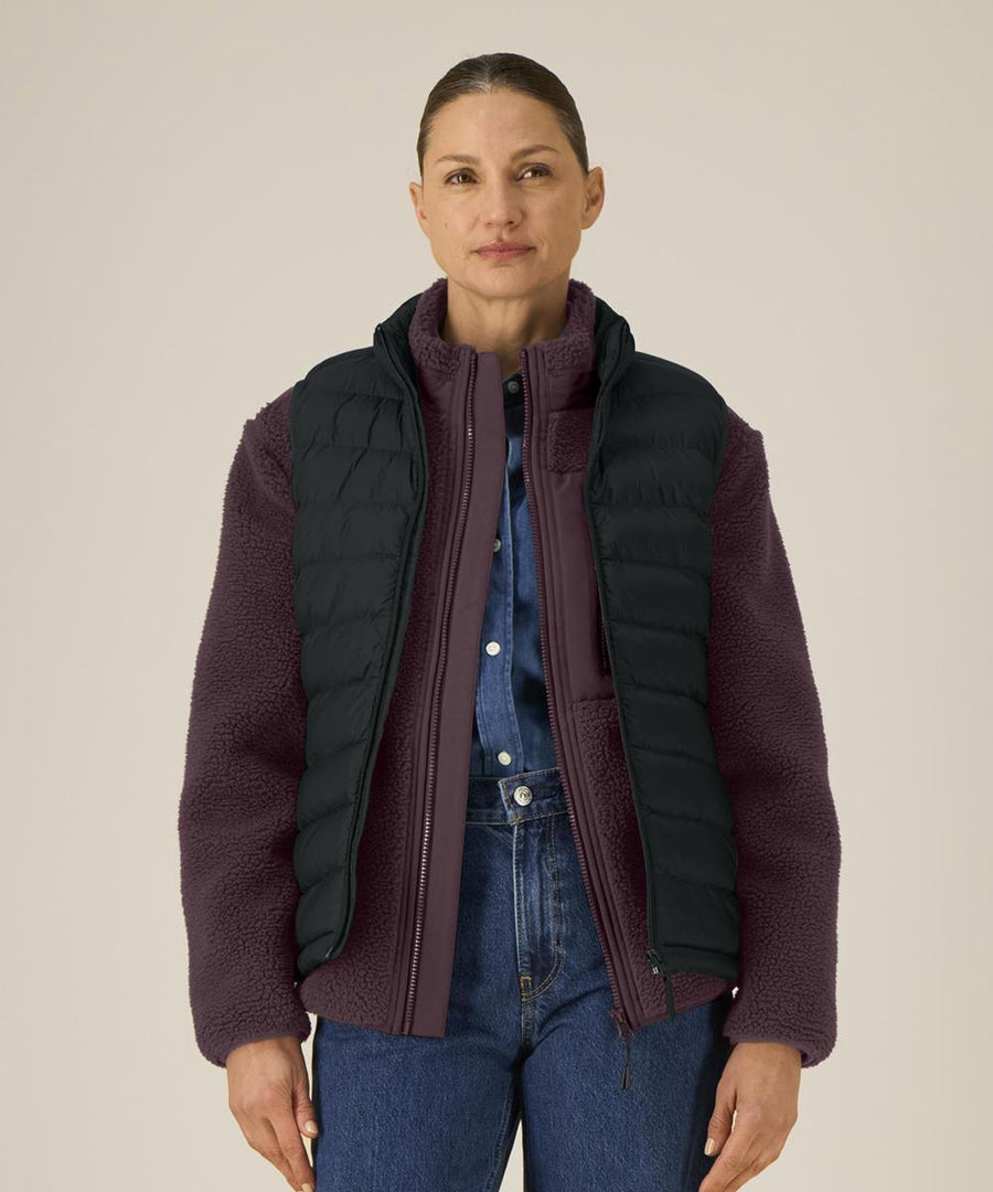 A woman with her hair tied back and a neutral expression faces the camera, wearing the Stanley/Stella Stella Climber 2.0 (STJW190) purple fleece jacket over a denim shirt and under a black padded gilet, against a plain light background.