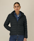 A person wearing the Stanley/Stella Stella Voyager 2.0 (STJW191) dark puffer jacket over a blue button-up shirt and blue jeans stands against a plain light background, one hand in their pocket, facing the camera with a neutral expression.
