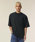 A person with short blonde hair stands against a plain light background, wearing the Stanley/Stella Blaster 2.0 (STTU959) loose black t-shirt and blue jeans, one hand in their pocket and a neutral expression.
