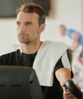 Man exercising on a stationary bike with a Towel City TC002 over his shoulder, looking forward.