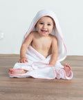 A smiling baby is sitting on a wooden floor, wrapped in a Towel City TC036 with a pink trim.