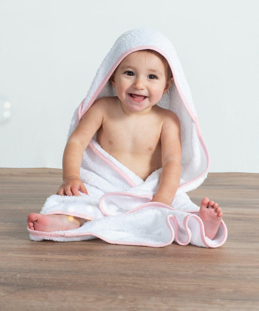 A smiling baby is sitting on a wooden floor, wrapped in a Towel City TC036 with a pink trim.