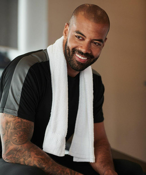 A man with a beard and a shaved head is sitting, wearing a black shirt with a white Towel City TC042 around his neck, smiling. He has tattoos on his right forearm.