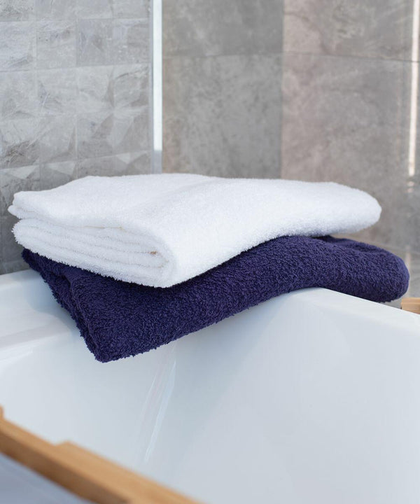 Two folded Towel City TC043 towels, one white and one dark blue, stacked on the edge of a bathtub against a tiled bathroom wall.