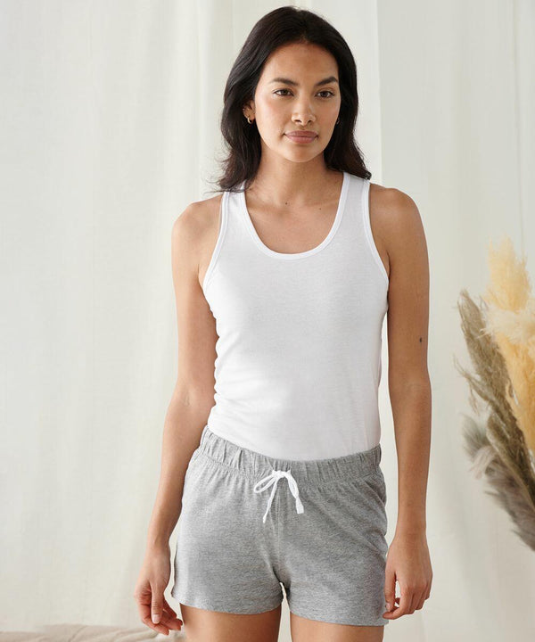 A person with long dark hair stands indoors wearing a white tank top and gray shorts from Towel City, looking at the camera, showcasing the TC052.