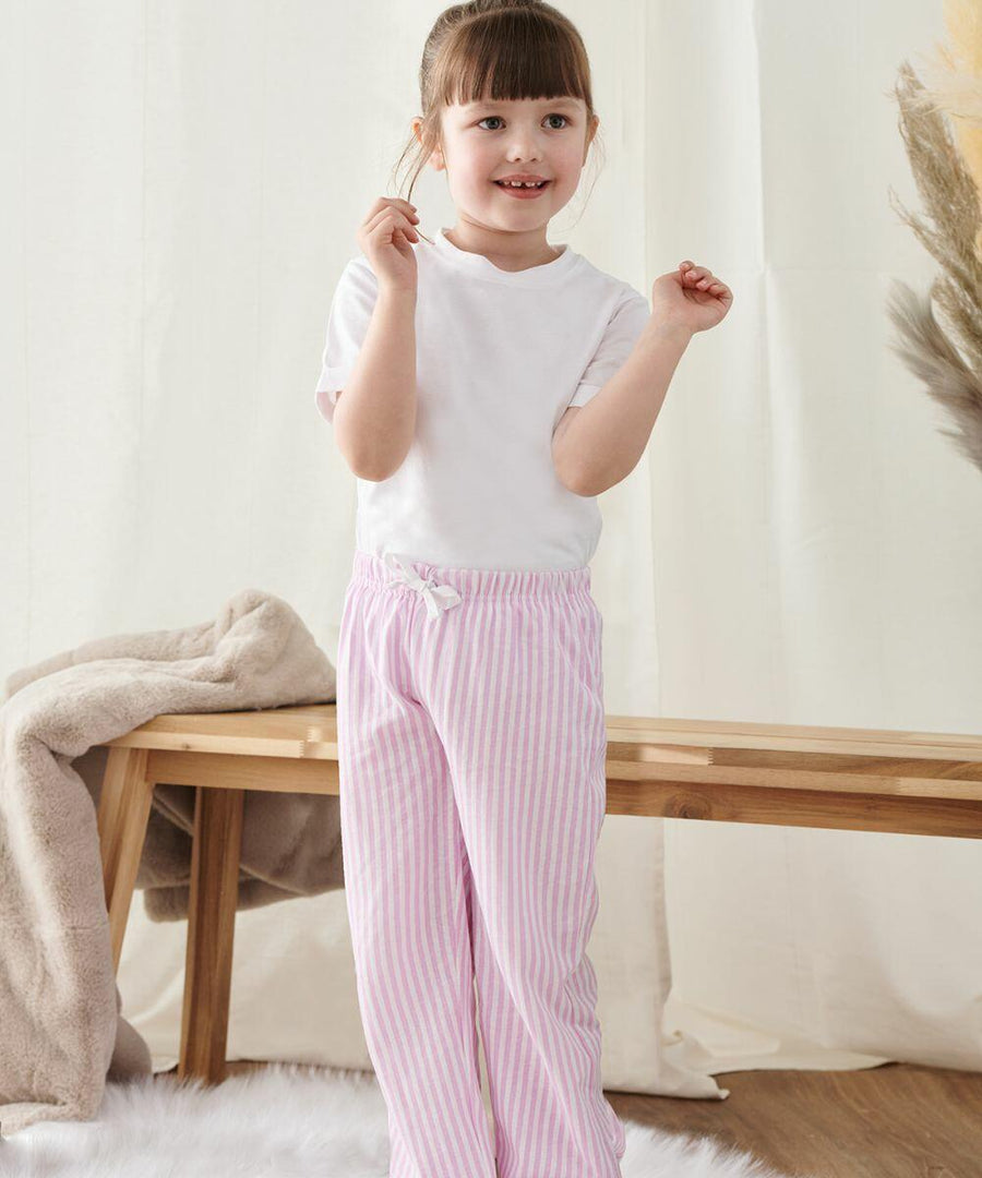 A child wearing a white shirt and pink striped pants stands indoors near a wooden bench, smiling and holding a small Towel City TC059 object in their hand.