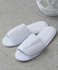 A pair of white TC067 open-toe slippers by Towel City placed on a gray tiled floor with a white wicker basket in the background.