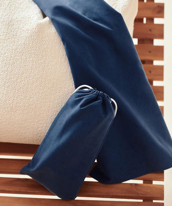 A Towel City quick-dry beach towel in dark blue is draped over the edge of a textured off-white mattress, with its matching fabric bag featuring a white drawstring resting on a wooden slatted surface nearby.