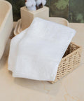 A neatly folded Organic guest towel with printable border by Towel City sits in a small rectangular woven basket on a beige worktop, with trays and cotton stems in the background.