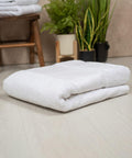 A neatly folded Towel City TC506 towel is placed on a light-colored wooden floor, with a wooden stool, folded towels, and potted plants in the background.