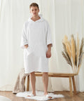 A man stands indoors wearing a white hooded robe and slippers. He is on a white rug next to a wooden bench with beige towels and decorative grass. Cream-colored curtains are in the background. The robe, identified as the TC810 from Towel City, adds an elegant touch to his appearance.