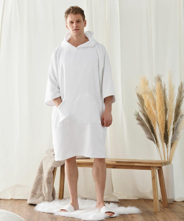 A man stands indoors wearing a white hooded robe and slippers. He is on a white rug next to a wooden bench with beige towels and decorative grass. Cream-colored curtains are in the background. The robe, identified as the TC810 from Towel City, adds an elegant touch to his appearance.