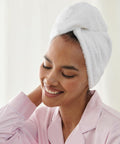 A person with a Towel City TC933 wrapped around their hair smiles contentedly. They are wearing a light pink pajama top and have their eyes closed.