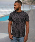 A man with a beard and short hair stands in an outdoor setting, wearing a black Colortone TD01M T-shirt and blue jeans. The background shows a railing and a body of water.