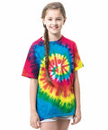 Girl with long braided hair wearing a colorful Colortone TD02B t-shirt and denim shorts, standing and smiling against a white background.