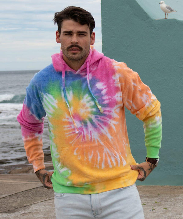 A man stands near the sea wearing a colorful Colortone TD31M hoodie and light jeans. A seagull is perched on a structure behind him.