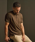 A man with short brown hair and a trimmed beard wears the Tee Jays Luxury Stretch Polo (1405) in brown and beige trousers, seated on a stool against a plain brown background, looking down with a neutral expression.