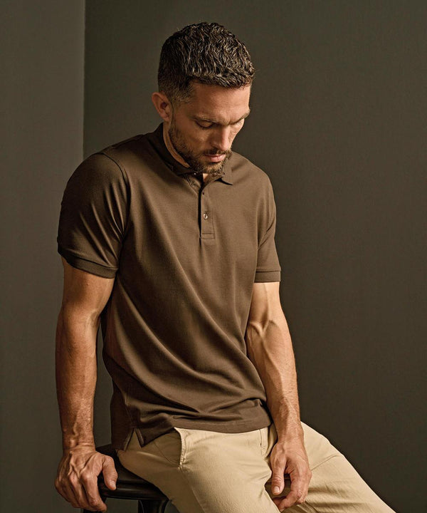 A man with short brown hair and a trimmed beard wears the Tee Jays Luxury Stretch Polo (1405) in brown and beige trousers, seated on a stool against a plain brown background, looking down with a neutral expression.