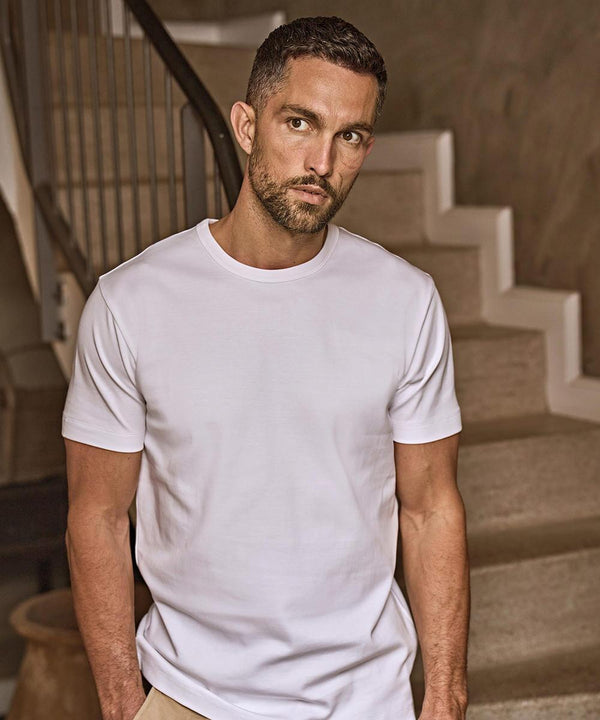 A man with short dark hair and a beard is wearing a plain white Tee Jays Interlock tee (520) and standing indoors in front of a staircase, looking directly at the camera.