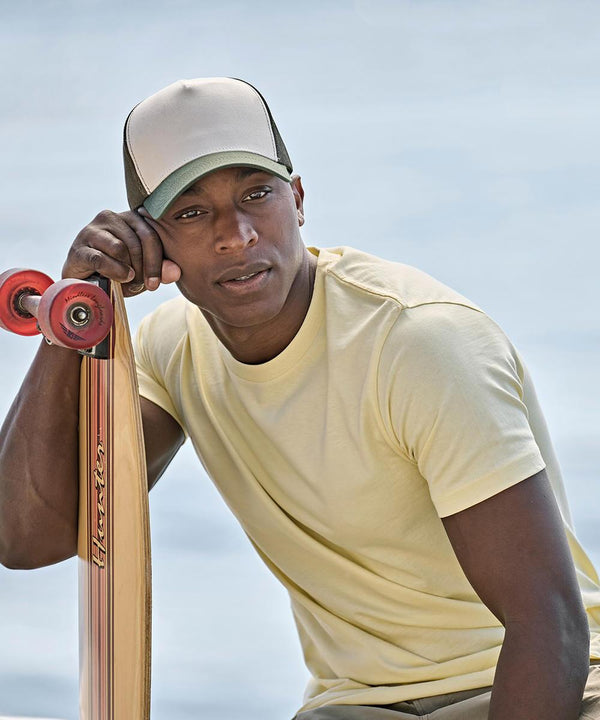 A person wearing the Tee Jays UNLABELLED luxury tee (1150) in light yellow and a beige cap leans on a longboard skateboard with red wheels, looking at the camera. The blurred outdoor background highlights the tee’s style.