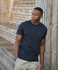 A man wearing a Tee Jays UNLABELLED tee (1140) in navy blue and beige shorts stands outdoors, leaning against a wooden wall with one hand in his pocket, looking to his left.