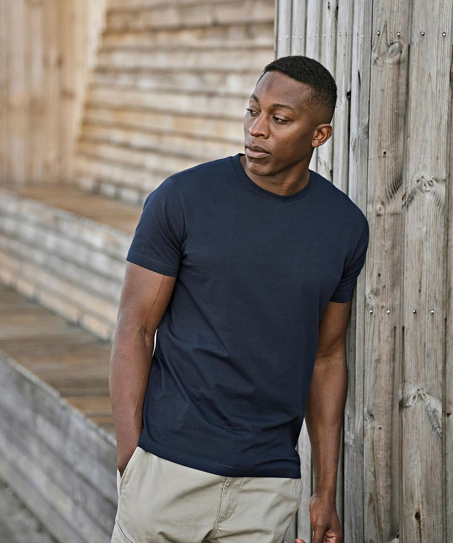 A man wearing a Tee Jays UNLABELLED tee (1140) in navy blue and beige shorts stands outdoors, leaning against a wooden wall with one hand in his pocket, looking to his left.