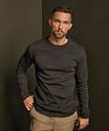 A man with short hair and a beard wears the Tee Jays Long Sleeve Interlock Tee (530) in black and beige trousers, standing against a dark background with one hand in his pocket, looking directly at the camera.