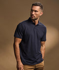 A man with short hair and a trimmed beard wears the Tee Jays Heavy polo (1400) in dark navy and tan trousers, standing against a plain brown background, looking slightly to the right.