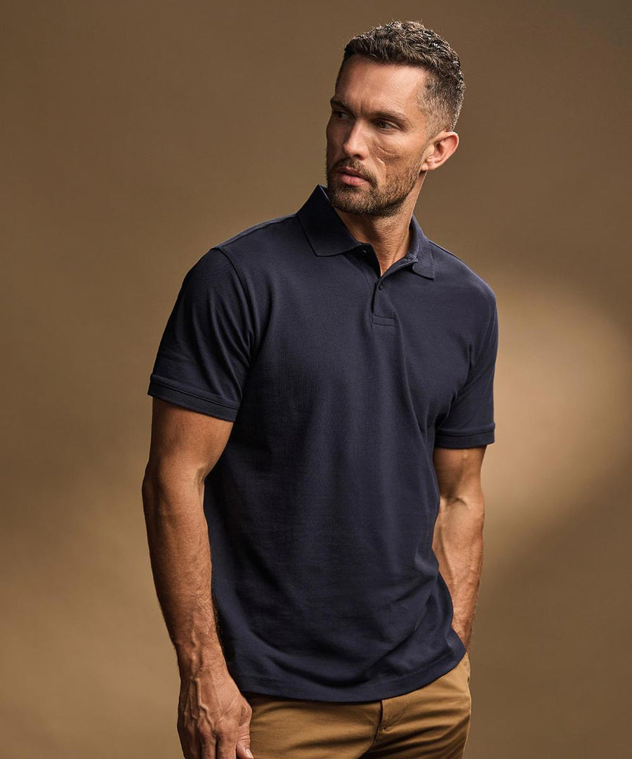 A man with short hair and a trimmed beard wears the Tee Jays Heavy polo (1400) in dark navy and tan trousers, standing against a plain brown background, looking slightly to the right.