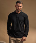 A man with short hair and a trimmed beard wears the Tee Jays Luxury Stretch Long Sleeve Polo (1406) in black with beige trousers, standing against a neutral brown background and showing a serious expression.