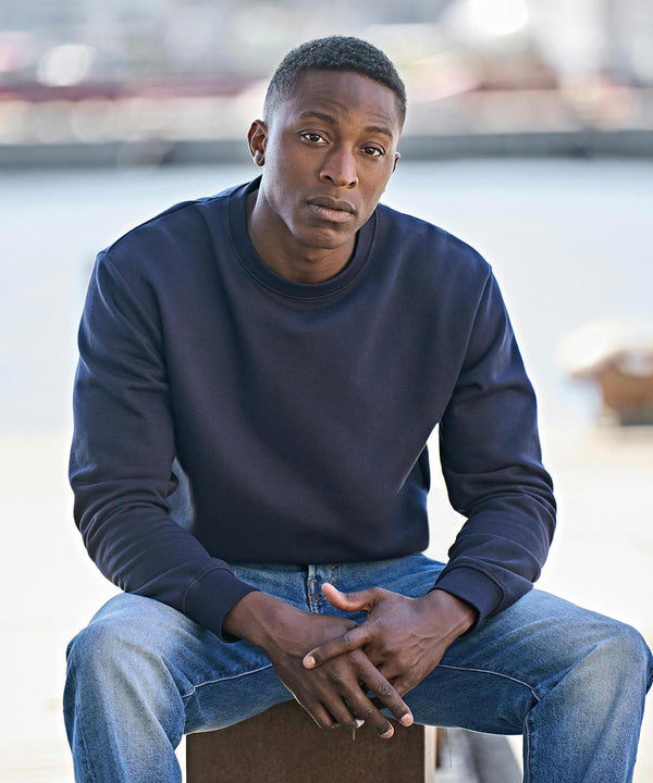 A man sits outdoors with a neutral expression, wearing the Tee Jays UNLABELLED sweatshirt (5150) in navy blue and blue jeans. His hands rest in his lap, and the background is blurred.