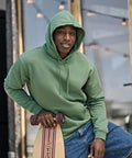 A person wearing a green Tee Jays UNLABELLED hooded sweatshirt (5152) sits outdoors, holding a longboard. The background features fairy lights and glass windows with a slight blur.