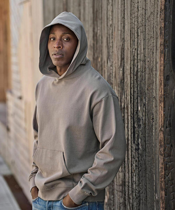A person stands outdoors in a Tee Jays UNLABELLED heavy loose-fit hooded sweatshirt (5162), hands in the front pocket, wearing blue jeans. They face the camera calmly beside a weathered wooden wall.