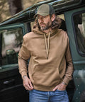 A man with long hair and a beard, wearing a Tee Jays Hooded Sweatshirt (5430), blue jeans, and a green lorry driver’s cap, stands by the open door of a green vehicle, looking to the side with one hand on his hip.