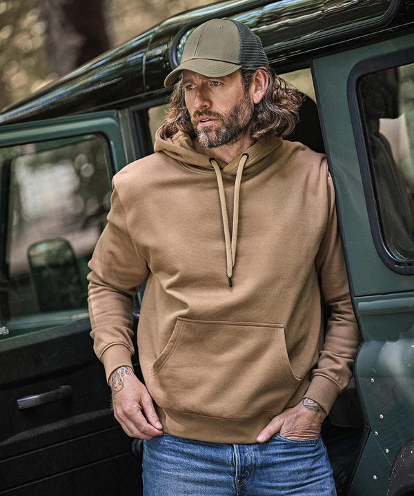 A man with long hair and a beard, wearing a Tee Jays Hooded Sweatshirt (5430), blue jeans, and a green lorry driver’s cap, stands by the open door of a green vehicle, looking to the side with one hand on his hip.