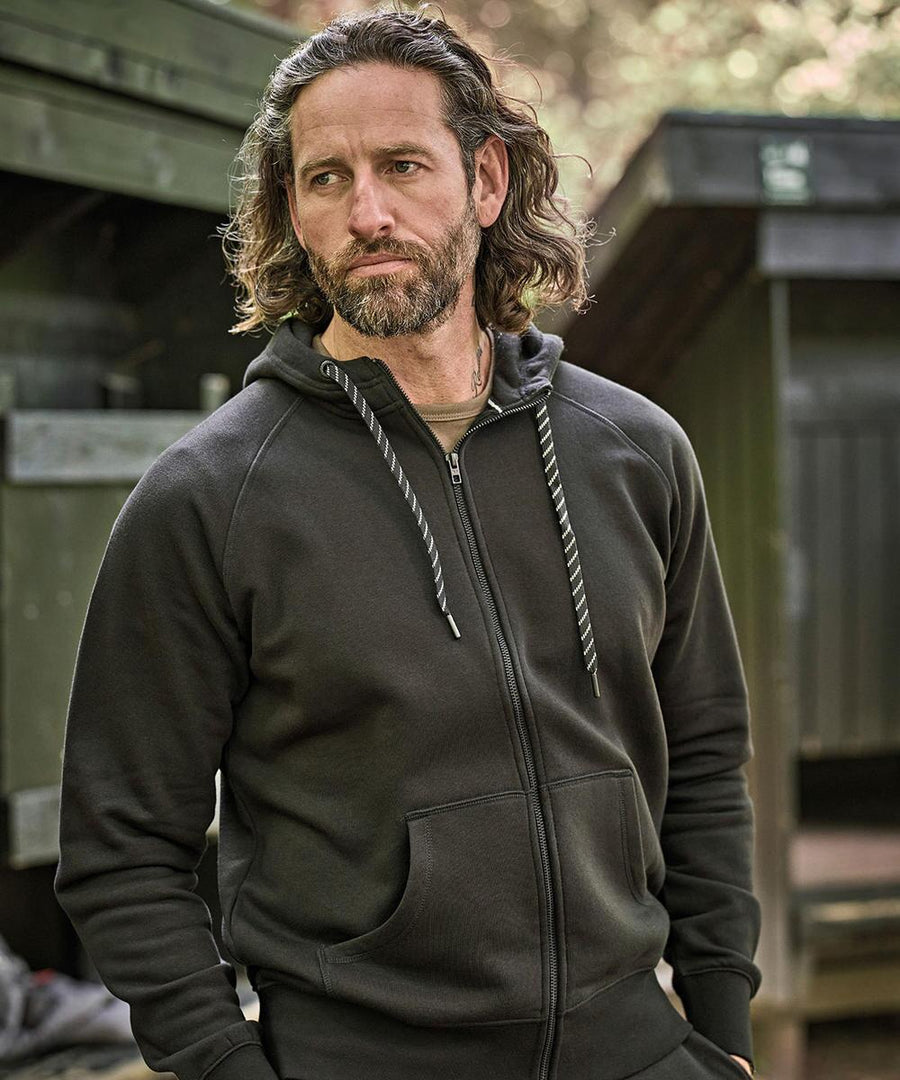 A man with long hair and a beard stands outdoors, wearing the Tee Jays Fashion full-zip hoodie (5435) featuring striped drawstrings and dark colour. He has his hands in the pockets, with green wooden structures in the background.