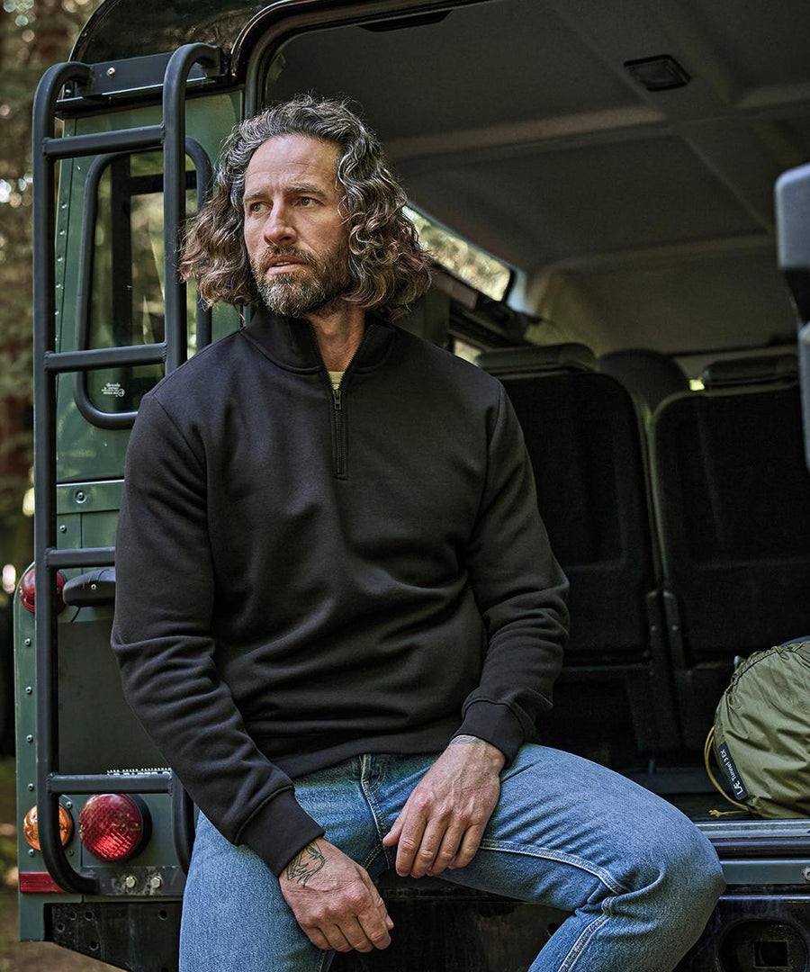 A man with shoulder-length wavy hair and a beard sits on the back of a vehicle wearing a black Tee Jays half-zip sweatshirt (5438) and blue jeans, with one hand on his knee and a green bag beside him.
